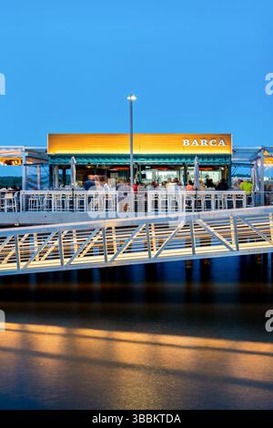 ALEXANDRIA, Virginie — Barca Pier & Wine Bar, un restaurant de tapas espagnoles, se trouve sur une jetée en bois qui s'étend dans le fleuve Potomac au bord de l'eau d'Alexandria au crépuscule. Le restaurant, qui a ouvert en 2021, présente un design en plein air avec des jeux de lumières éclairant le coin salon extérieur à l'approche de la soirée. Les convives apprécieront les petites assiettes et boissons d'inspiration espagnole tout en admirant la vue sur le fleuve Potomac et le front de mer d'Alexandria, avec la lueur chaude du coucher du soleil créant une atmosphère pittoresque. Banque D'Images