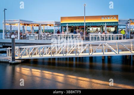 ALEXANDRIA, Virginie — Barca Pier & Wine Bar, un restaurant de tapas espagnoles, se trouve sur une jetée en bois qui s'étend dans le fleuve Potomac au bord de l'eau d'Alexandria au crépuscule. Le restaurant, qui a ouvert en 2021, présente un design en plein air avec des jeux de lumières éclairant le coin salon extérieur à l'approche de la soirée. Les convives apprécieront les petites assiettes et boissons d'inspiration espagnole tout en admirant la vue sur le fleuve Potomac et le front de mer d'Alexandria, avec la lueur chaude du coucher du soleil créant une atmosphère pittoresque. Banque D'Images