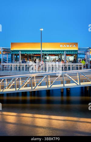 ALEXANDRIA, Virginie — Barca Pier & Wine Bar, un restaurant de tapas espagnoles, se trouve sur une jetée en bois qui s'étend dans le fleuve Potomac au bord de l'eau d'Alexandria au crépuscule. Le restaurant, qui a ouvert en 2021, présente un design en plein air avec des jeux de lumières éclairant le coin salon extérieur à l'approche de la soirée. Les convives apprécieront les petites assiettes et boissons d'inspiration espagnole tout en admirant la vue sur le fleuve Potomac et le front de mer d'Alexandria, avec la lueur chaude du coucher du soleil créant une atmosphère pittoresque. Banque D'Images