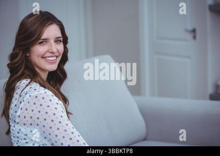 Femme souriante assise sur un canapé rembourré gris clair dans le salon, soulignant un décor minimal Banque D'Images