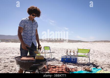 Griller homme tournant le maïs et les saucisses avec des pinces en métal sur la plage de sable, avec glacière et chaises Banque D'Images