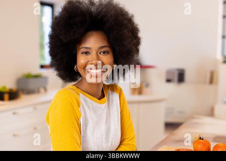 Femme afro-américaine appuyée sur l'îlot de cuisine, souriante et présentant des tomates orange dans la cuisine Banque D'Images