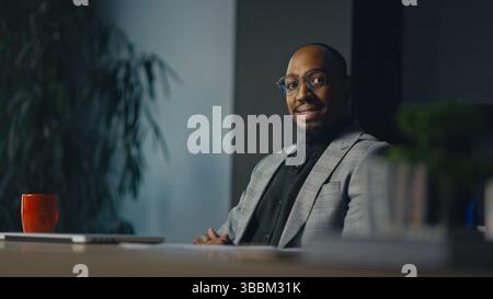 Homme d'affaires confiant assis au bureau, souriant à la caméra dans une posture détendue, vêtu d'un costume à carreaux moderne. Concept de leadership Banque D'Images