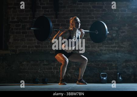 Femme en position squat tient la barre sur les épaules, se préparant à l'ascenseur dans le gymnase sous l'éclairage moody avec fond de mur de briques, montrant la force et Banque D'Images