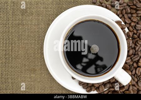 Une tasse de café noir fumant se trouve sur une soucoupe blanche, entourée de grains de café éparpillés sur la surface en toile de jute. la réflexion dans le café ajoute une touche d'intrig Banque D'Images