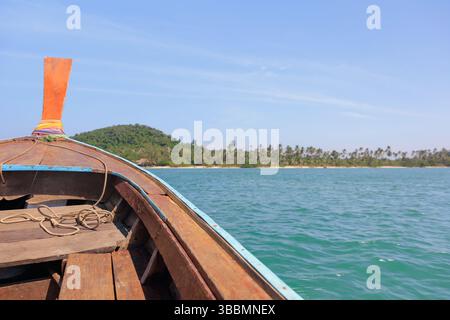 Proue d'un bateau à longue queue en bois naviguant sur de belles eaux turquoises claires sous un ciel bleu vif, avec une île vallonnée verte au loin. Banque D'Images