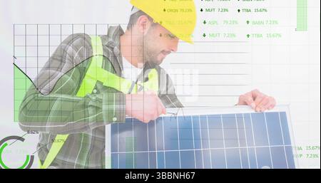 Homme portant un casque jaune, gilet de sécurité ajustant le panneau solaire en studio, avec des graphiques financiers Banque D'Images