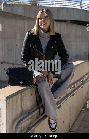 Femme à la mode portant un Jean déchiré et une veste en cuir se trouve sur un mur de béton dans un environnement urbain moderne Banque D'Images
