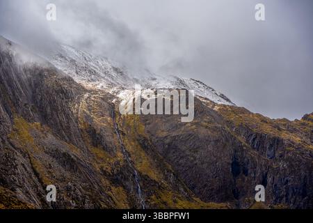 Les rochers poussiéreux de neige de l’amphithéâtre glaciaire de Cwm Idwal se révèlent dans des détails ornés de brume avec une fine cascade traçant d’anciennes strates rocheuses Banque D'Images