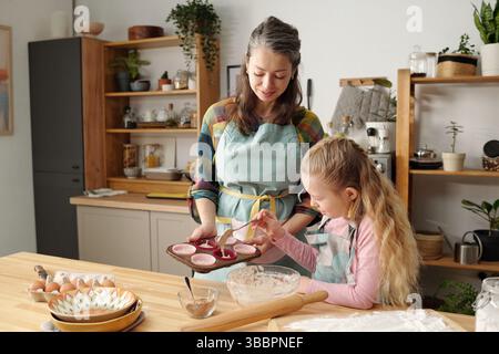 Mère et fille souriantes appréciant de cuisiner ensemble dans une cuisine chaleureusement éclairée, portant des tabliers et utilisant des ustensiles de cuisine sur un comptoir en bois avec divers ingrédients placés autour d'eux Banque D'Images
