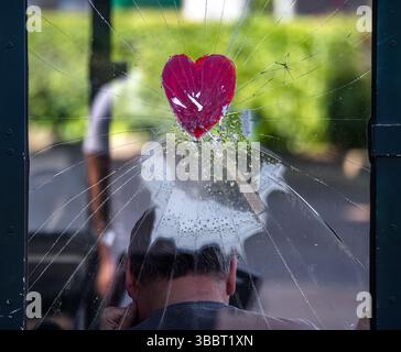 Une fenêtre en verre fissurée avec un autocollant cœur rouge au centre, montrant une figure floue assise derrière elle. L'arrière-plan présente de la verdure et des conseils Banque D'Images