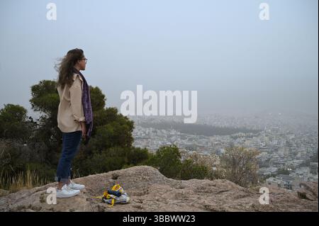 Athènes, Grèce, 16 mai 2025. Une femme regarde la ville tandis qu'Athènes est couverte de nuages de poussière. Une épaisse couverture de poussière saharienne a balayé la capitale grecque vendredi, jetant de la brume sur la ville et suscitant des avertissements sanitaires de la part des autorités. Crédit : Dimitris Aspiotis/Alamy Banque D'Images