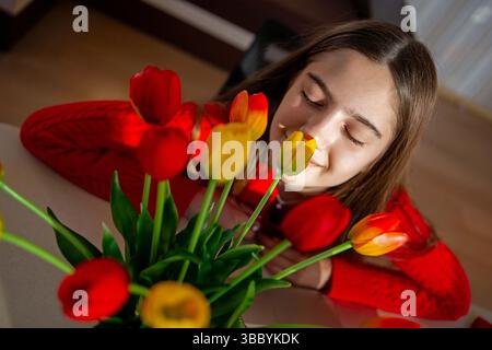 Femme appréciant le parfum des tulipes vibrantes. Une jeune femme dans un pull rouge sourit paisiblement les yeux fermés alors qu'elle se penche vers un bouquet coloré Banque D'Images