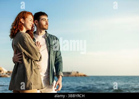 Jeune couple debout côte à côte près de l'océan, embrassant doucement tout en regardant devant, baigné dans la lumière chaude du soleil lors d'un après-midi côtier paisible. La vie Banque D'Images