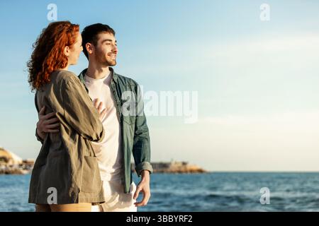 Jeune couple profite d'un moment romantique au bord de l'océan, debout bras dans bras avec des expressions joyeuses alors qu'ils regardent la mer calme sous le coucher du soleil chaud lig Banque D'Images