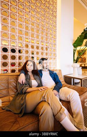 Un jeune couple détendu se prélasse ensemble sur un canapé moderne en cuir dans un hall lumineux à la décoration contemporaine et aux murs géométriques. Style de vie. Banque D'Images
