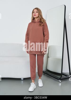 Femme portant un sweat à capuche et un pantalon de couleur rouille se tient devant un grand miroir, les mains à ses côtés, des baskets blanches aux pieds, à côté d'une lumière Banque D'Images