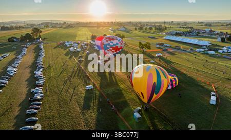 Des montgolfières aux couleurs vives sont gonflées dans un vaste champ tôt le matin. Les véhicules sont garés à proximité car la lumière du soleil baigne le paysage dans des tons chauds. Banque D'Images
