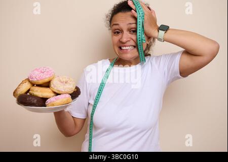 Gros plan d'une femme tenant une assiette de beignets colorés tout en montrant une expression conflictuelle, symbolisant la santé et les choix d'indulgence. Banque D'Images