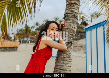Une femme en robe rouge est appuyée contre un arbre sur une plage. La scène est sereine et paisible, avec la femme posant pour une photo Banque D'Images