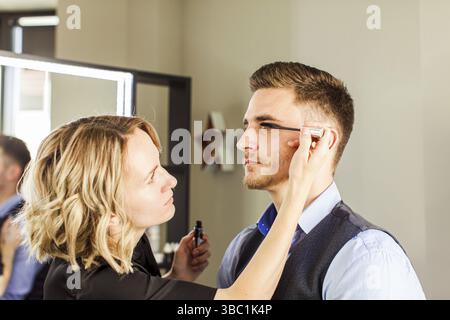 Préparation professionnelle des acteurs avant la prise de vue. Beau jeune homme appliquant par la forme professionnelle de visagiste. Maquillage pour hommes Banque D'Images