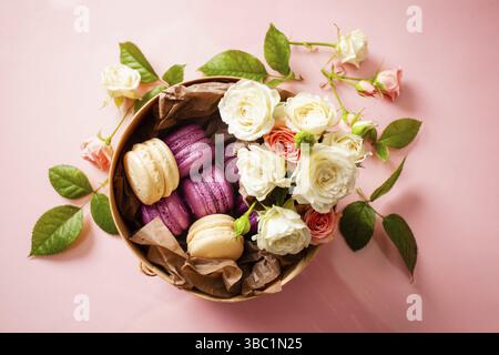 Vue de dessus d'une boîte ronde remplie de délicieux macarons blancs et violets, décorés de roses. Boîte cadeau Sweet. Fond rose Banque D'Images
