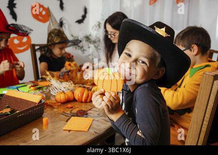 Portrait d'un adorable garçon souriant dans un chapeau de cow-boy portant une citrouille en papier faite à la main. L'écolier démontre ce qu'il a créé lors de l'atelier d'halloween. Créatif Banque D'Images