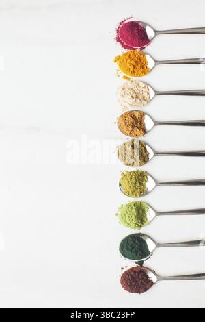 Divers types de poudres superalimentaires colorées en cuillères métalliques sur table blanche, espace de copie Banque D'Images