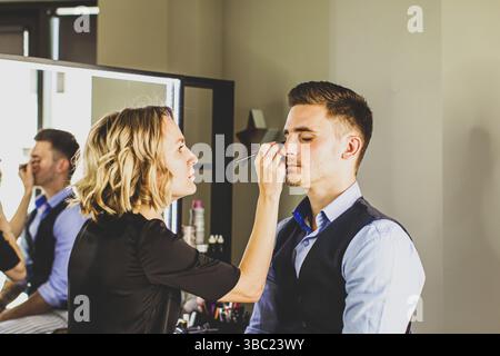 Maquilleuse faisant du maquillage professionnel pour jeune homme. Bel homme mannequin en studio Banque D'Images