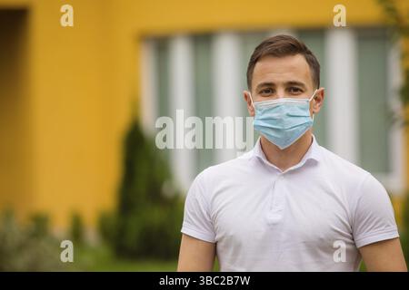 Portrait d'un jeune homme portant un t-shirt blanc et un masque médical. Il se tient à l'extérieur et regarde la caméra Banque D'Images
