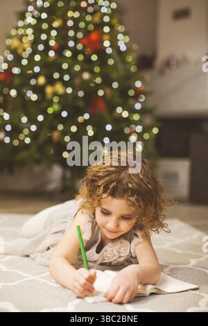 Des vacances de Noël magiques et un concept d'enfance heureux. Fille souriante faisant la liste de souhaits ou la lettre au Père Noël à la maison. Sapin de Noël sur fond, flou Banque D'Images