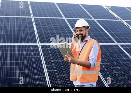 Le jeune électricien dans un gilet et un casque tient une tablette et parle au téléphone. Il se dresse sur fond de panneaux solaires Banque D'Images