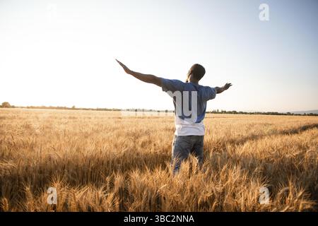 Vue arrière jeune homme méconnaissable tenant les mains vers le haut, saluant le soleil, debout dans le champ de blé.Un jeune homme se précipitant, passant du temps à cou Banque D'Images