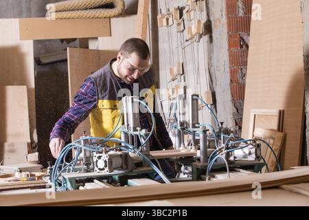 Portrait d'un jeune employé gai travaillant dans le bois utilisant une machine pneumatique dans un atelier poussiéreux. Production de meubles. Homme dans des lunettes de protection Banque D'Images