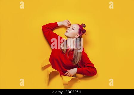 Belle jeune femme avec des petits pains de couleur rose, maquillage brillant, portant le sweat à capuche rouge s'étirant après le sommeil, regardant hors de déchiré dans le papier jaune wal Banque D'Images