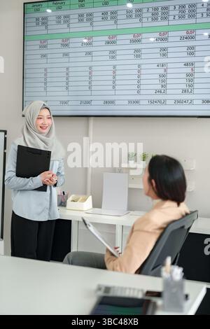 Deux femmes d'affaires dans un bureau analysant des données financières sur un écran. Une femme asiatique en hijab présente des données à un collègue. Discuter des tendances, des ventes, p Banque D'Images