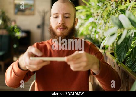 Homme roux barbu scannant le dépôt de chèque à distance tout en utilisant le téléphone portable Banque D'Images