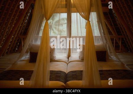 Un lit double avec des rideaux à baldaquin transparents, des oreillers moelleux et un éclairage chaleureux dans un intérieur de cabane en bambou Banque D'Images