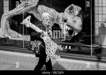 Femme portant une girafe en peluche marche devant la grande figure Tyrannosaurus Rex dans la vitrine du magasin. Regent Street, Londres, Royaume-Uni. Banque D'Images