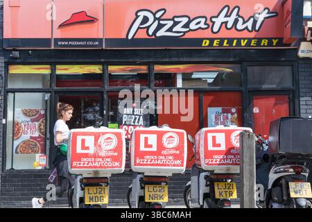 Une femme entre dans un magasin de plats à emporter Pizza Hut car plusieurs motos équipées de boîtes chauffantes de livraison de nourriture et de plaques en « l » sont garées près de la porte d'entrée au Royaume-Uni Banque D'Images