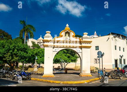 Arche ornementale de style colonial se trouve en bonne place dans le Parque de la Merced dans le centre de Leon, Nicaragua. Encadrée par des motos, des palmiers et des bancs, la structure évoque le riche patrimoine architectural de la ville sous un ciel bleu vif Banque D'Images