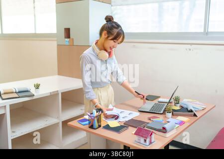 Jeune femme asiatique designer d'intérieur travaille à son bureau sur un ordinateur portable entouré d'échantillons de matériaux de modèles de maison et de croquis de design d'intérieur dans sa créa Banque D'Images
