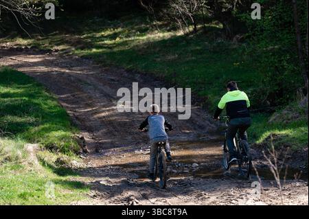 Deux personnages sur des vélos traversent un chemin boueux dans un cadre boisé. L'individu le plus âgé dirige le plus jeune, créant une scène réconfortante de o partagé Banque D'Images