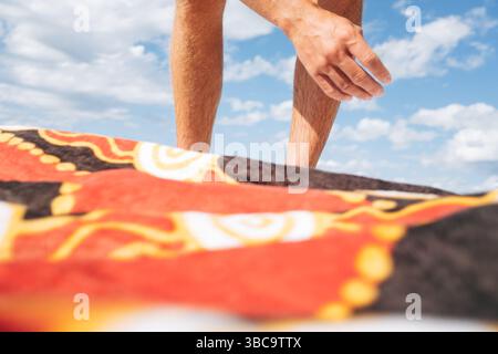Homme atteignant au-dessus d'une serviette de plage à motifs sous un ciel bleu vif Banque D'Images