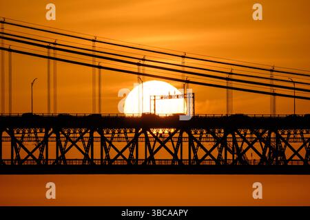Vue rapprochée du coucher de soleil directement derrière le pont 25 de Abril à Lisbonne, Portugal, ciel orange chaud avec quelques nuages, voitures et câbles en silhouette contre le soleil incandescent, scène de coucher de soleil urbain saisissant, véhicules traversant le pont, brume de chaleur estivale, soleil brillant Banque D'Images