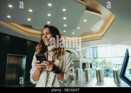 Une femme d'affaires indienne confiante sourit tout en tenant un smartphone dans un environnement de bureau moderne et élégant. L'espace lumineux met en valeur son pr Banque D'Images