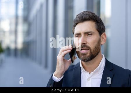 Un bel homme d'affaires, portant un costume, utilise son téléphone en se tenant devant un immeuble de bureaux, arrière-plan flou. Banque D'Images