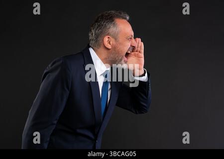 Homme d'âge moyen en costume bleu marine criant latéralement avec sa main à la bouche, montrant l'urgence ou la colère, sur un fond noir. Banque D'Images