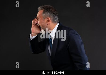 Homme mûr dans un costume criant en avant avec ses mains creusées autour de sa bouche, exprimant l'urgence, l'annonce ou la protestation, sur un fond sombre. Banque D'Images