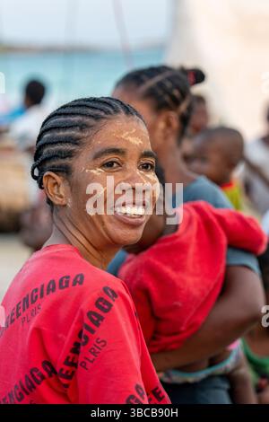 Une femme souriante dans une chemise rouge avec un masque facial traditionnel lors des courses de canoë à Andavadoaka, Madagascar. Elle rayonne de joie, reflétant le vibra Banque D'Images